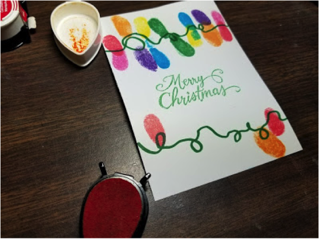 The sentiment "Merry Christmas" is stamped in green between two images of christmas light strings created with multi-colored inked fingerprints and markers.